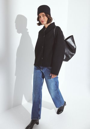 Svart ullknappad jacka, blå jeans och svarta klackstövlar. En svart väska slängd över axeln. Modell som bär en matchande svart mössa.