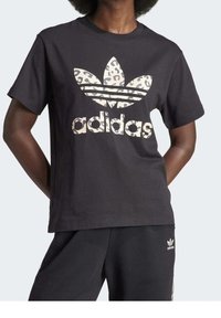 Person som bär en svart Adidas-t-shirt med leopardmönster på logotypen och matchande svarta Adidas-byxor med en liten vit logotyp nära fickan.