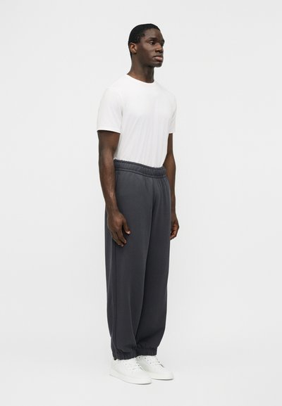 Άνδρας που στέκεται φορώντας λευκό κοντομάνικο πουκάμισο, σκούρο γκρι παντελόνι με ελαστική μέση και λευκά αθλητικά παπούτσια, μπροστά σε απλό ανοιχτό φόντο.