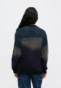 Asmuo su ilgais juodais plaukais, vilkintis pūkuotą, spalvotą megztinį jūrų mėlynos, pilkos ir juodos spalvų deriniu, derinamas su mėlynomis krovinių kelnėmis, atsisukęs atgal.