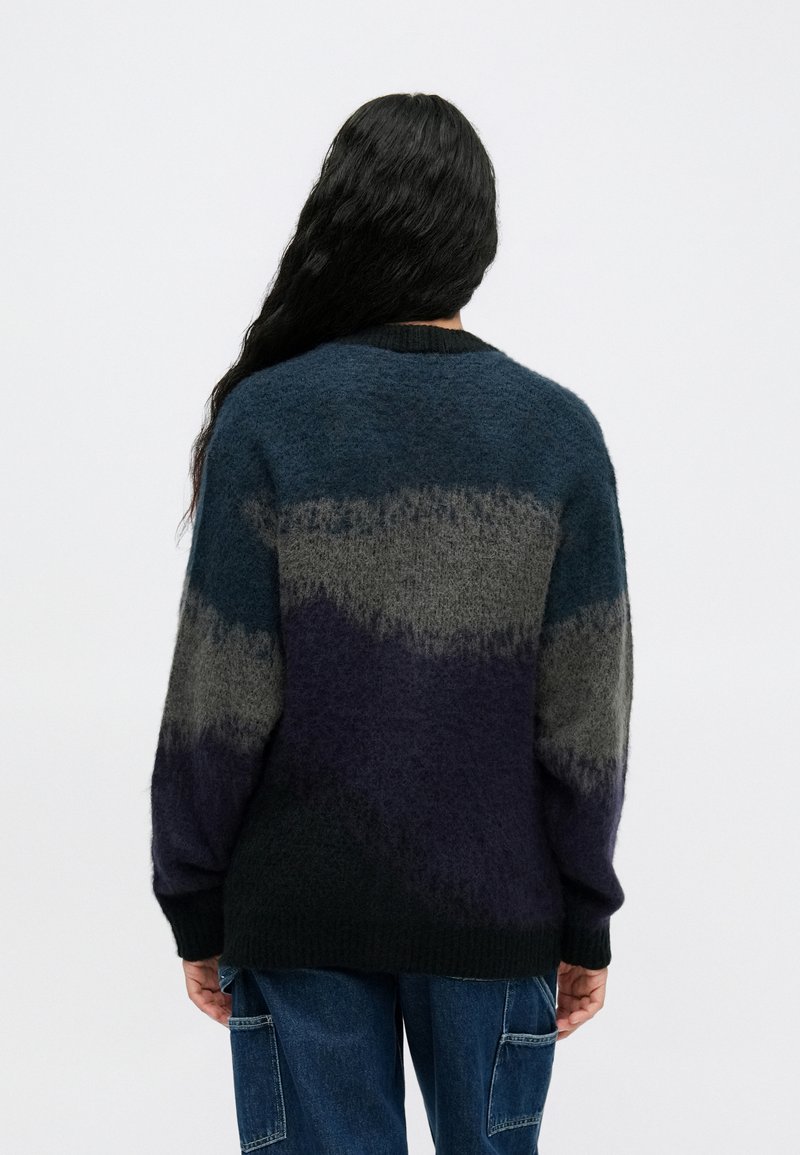 Person with long black hair wearing a fuzzy, color-blocked sweater in navy, gray, and black, paired with blue cargo jeans, facing away.