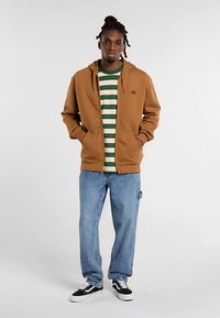 Brun hættejakke med lynlås over en grøn og hvidstribet t-shirt, kombineret med løse blå jeans og sorte sneakers, med et logo på hættejakken.