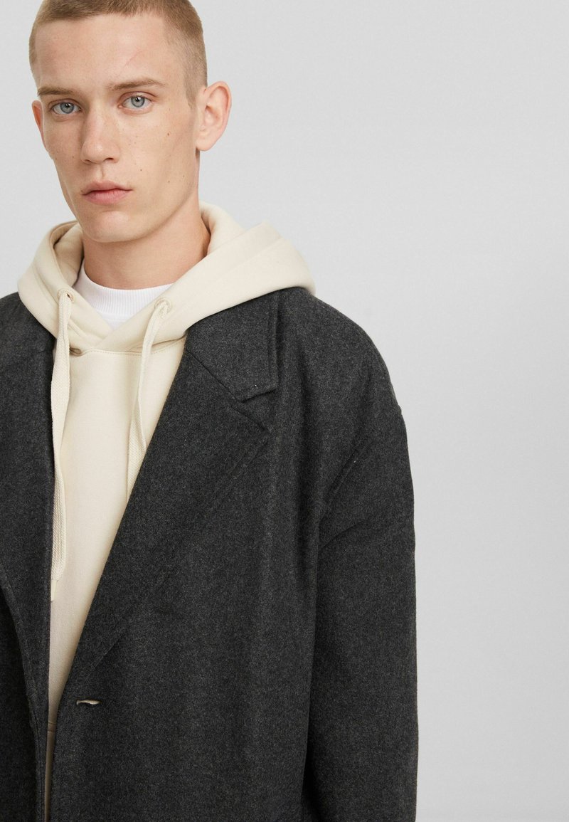 Jonge man met kort licht haar, gekleed in een roomkleurige hoodie onder een donkergrijze oversized wollen jas, met een neutrale uitdrukking, op een lichtgrijze achtergrond.