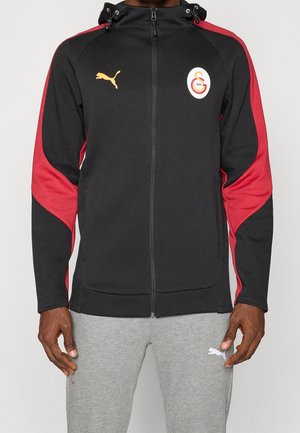 Sweat à capuche noir zippé avec des bandes rouges sur les manches, logo Puma jaune, logo Galatasaray sur la poitrine, porté avec un pantalon de survêtement Puma gris clair.