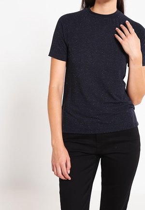 Femme portant un t-shirt uni bleu marine foncé à manches courtes et un pantalon noir, debout avec une main reposant près de sa clavicule.