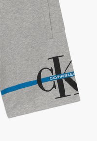 Šedé bavlněné šortky s velkým černým logem "CK" a textem "CALVIN KLEIN JEANS" modrou barvou přes přední část, s bočními kapsami.