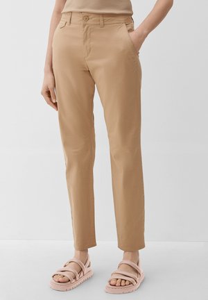 Lower body of a person wearing beige straight-leg pants and light pink padded sandals, standing with one hand in pocket against a plain background.