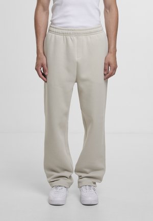 Persona che indossa pantaloni beige larghi da ginnastica e sneakers bianche, in piedi contro uno sfondo chiaro e uniforme.