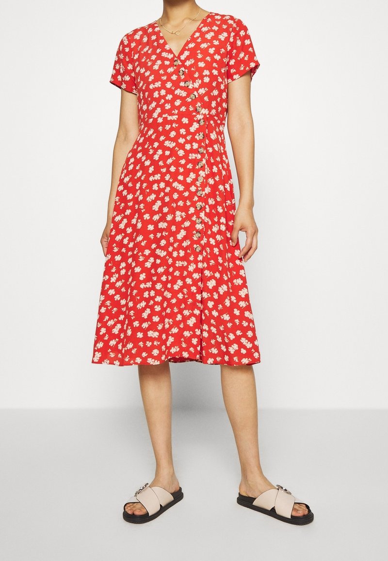 Vestido envuelto rojo con estampado floral, mangas cortas, escote en V y detalle de botones en el frente. Combinado con sandalias ligeras tipo slip-on.