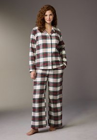 Woman with curly red hair wearing white, red, green, and blue plaid long-sleeve pajama set, standing barefoot against a gray background.