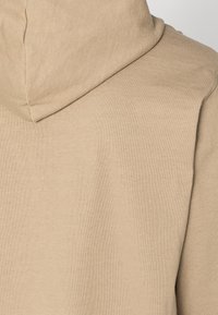 Beige hoodie med en slät textur, som har en dragsko i huvan och ribbad sömnad längs kanterna. Minimalistisk design utan mönster.