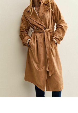 Femme portant un long trench-coat marron à double boutonnage avec une ceinture nouée à la taille et les mains dans les poches sur un fond uni.