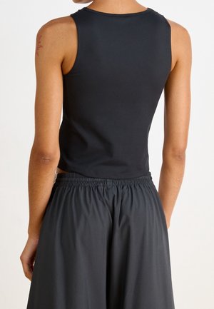 Personne portant un top court noir sans manches et un pantalon large noir, vue de dos sur un fond blanc uni.