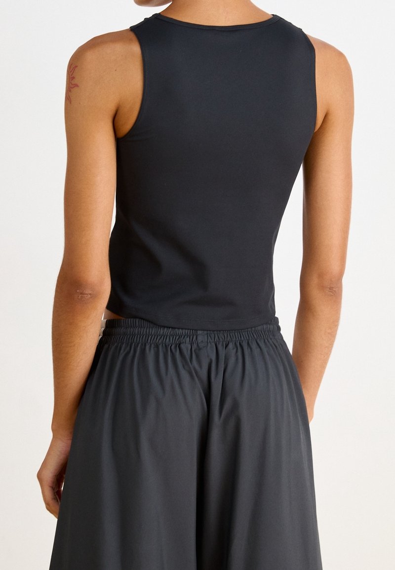 Persona vistiendo un top corto sin mangas negro y pantalones negros holgados, mostrada de espaldas sobre un fondo blanco liso.