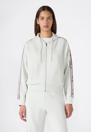 Joven mujer vistiendo una sudadera blanca con cremallera y rayas de logo en las mangas, y pantalones blancos a juego, de pie frente a un fondo blanco liso.