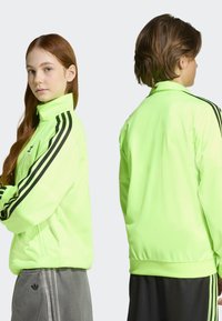 Neongrön zip-up jacka med svarta ränder, hög krage och ribbade muddar, visad fram- och bakifrån. Slät yta, atletisk design.