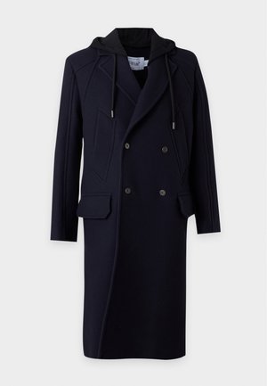 Homme portant un long manteau noir croisé avec capuche, t-shirt blanc, jean bleu et baskets blanches, debout devant un fond uni.