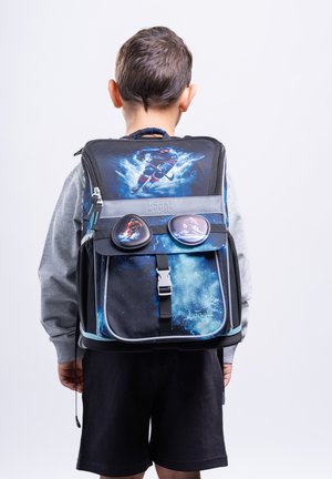 Boy wearing a black and blue backpack with a hockey player design and two matching badge pins, standing against a plain light background.