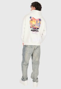 Person iført hvid hættetrøje med farverig solnedgang, palmetræer og sportsvogns grafik, sammen med slidte blå jeans og hvide sneakers.