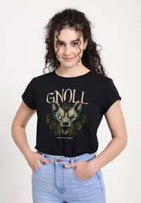 Černé bavlněné tričko s grafikou vrčícího gnolla v zelené a černé barvě s textem "GNOLL" nahoře, nošené s světle modrými džíny.