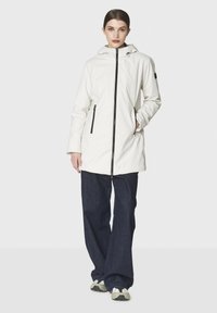 Nainen seisoo päällään valkoinen hupullinen takki, jossa on mustat vetskarit, tummansiniset leveälahkeiset farkut ja valkoiset tennarit.