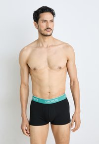 Boxer-shorts noirs avec une taille teal arborant le logo "LACOSTE". Le tissu semble doux et lisse avec un design ajusté.