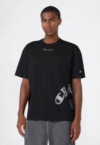 Man die staat, gekleed in een zwart Champion t-shirt met logo op de borst, mouw en onderzijde, gecombineerd met grijze shorts tegen een effen achtergrond.