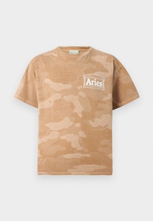 Tânăr cu păr creț stă privind spre dreapta, purtând un tricou camuflat maro deschis cu logo-ul "Aries", pantaloni largi verde închis și adidași alb-negru.
