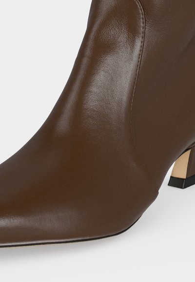 Gros plan d'une bottine à talon en cuir marron avec un bout pointu et un talon carré sur fond blanc.
