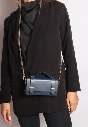 Navy blue leather handbag with gold hardware accents, boxy design, top handle, and chain strap. Worn against a black blazer.