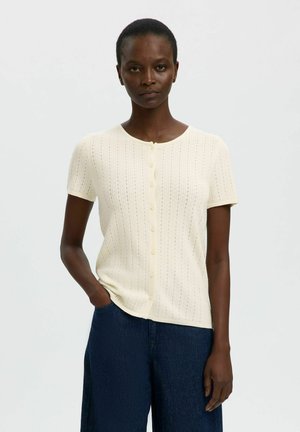 Vrouw staand tegen een effen achtergrond, draagt een crèmekleurige gebreide knoopblouse met korte mouwen en donkerblauwe broek, één hand in de zak.