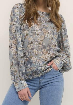 Vrouw die een doorschijnende, lange mouwen bloemige blouse in gedempte tinten draagt, gecombineerd met lichtblauwe spijkerbroek, met één hand in de zak, staand tegen een effen achtergrond.