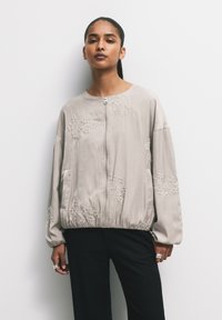 Giubbotto bomber beige con ricami floreali, materiale leggero, chiusura con zip, orlo e polsini elasticizzati, e tasche laterali.
