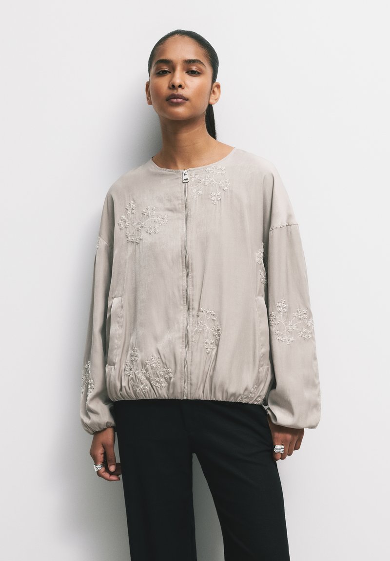 Giubbotto bomber beige con ricami floreali, materiale leggero, chiusura con zip, orlo e polsini elasticizzati, e tasche laterali.