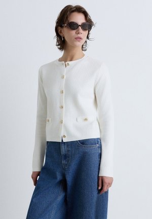 Woman wearing black sunglasses, white buttoned knit cardigan, and loose blue jeans against plain light background.