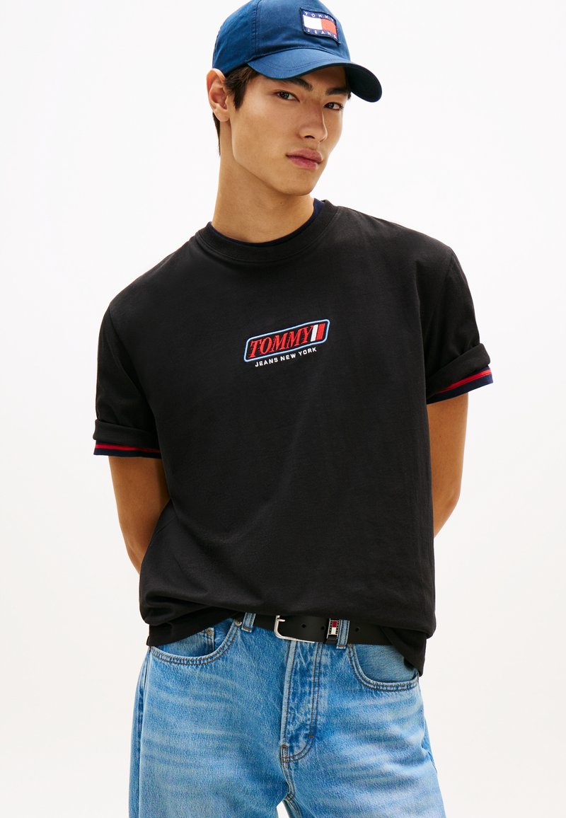 Νέος άνδρας που φοράει μαύρο t-shirt Tommy Jeans, μπλε καπέλο και ανοιχτό μπλε τζιν, στέκεται με τα χέρια πίσω από την πλάτη.