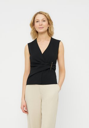 Femme aux cheveux blonds portant un haut cache-cœur sans manches noir avec une épingle et un pantalon beige, debout avec une main dans la poche, sur fond blanc.