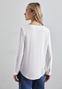 Weißes Langarmblusenshirt mit Rundhalsausschnitt und Seitenschlitzen. Leichter, glatter Stoff mit abgerundetem Saum, kombiniert mit blauen Denimjeans.