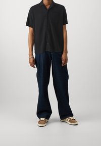 Uomo che indossa una camicia nera a maniche corte con bottoni, jeans larghi blu scuro, sneakers marroni e bianche, e un braccialetto argentato sul polso sinistro.