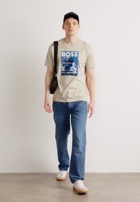 Tricou bej cu un imprimeu grafic albastru, care prezintă "BOSS", asortat cu blugi albaștri deschiși și adidași albi. Include o șapcă neagră și o geantă de umăr.