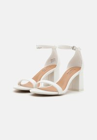 Sandales blanches à talons avec une bride autour de la cheville, un design à bout ouvert, un talon carré et une finition synthétique lisse. L'intérieur est beige avec des coutures décontractées.