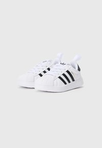 Λευκά αθλητικά παπούτσια Adidas Superstar με μαύρες ρίγες, σχεδιασμό με προστασία δαχτύλων, λευκά κορδόνια και ταινίες έλξης στο πίσω μέρος, απεικονιζόμενα σε απλό φόντο.