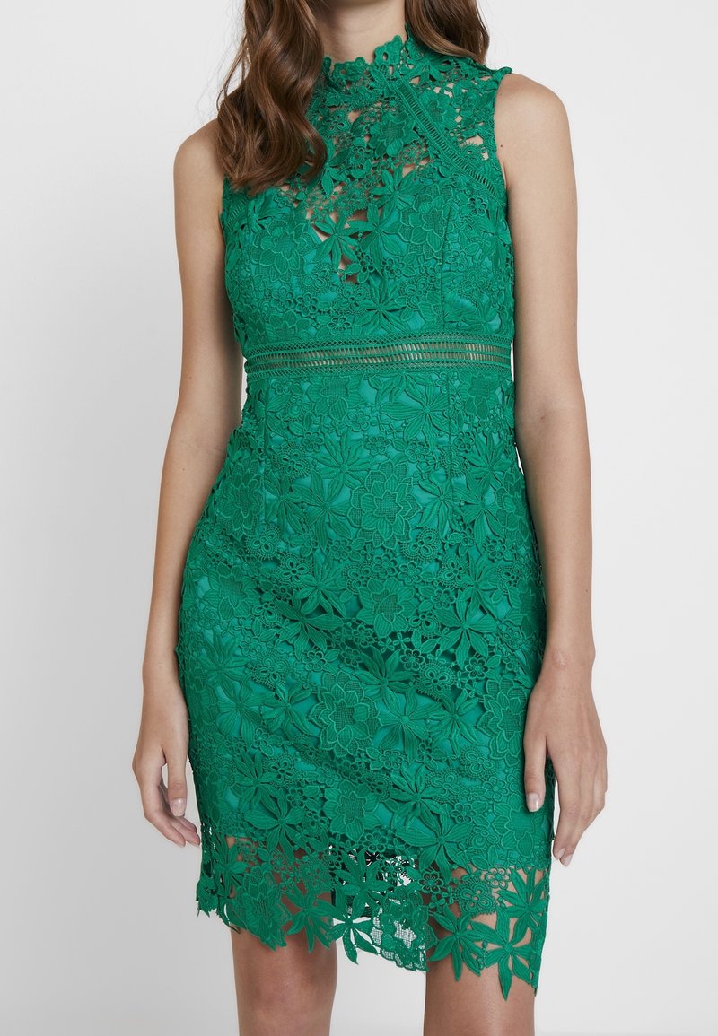 Robe en dentelle verte avec des motifs floraux, col haut, sans manches et ourlet festonné. Le matériau est délicatement texturé.