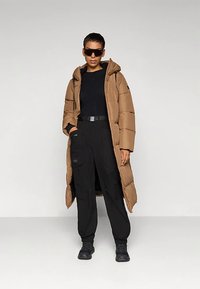 Giacca imbottita marrone con cappuccio, caratterizzata da un design trapuntato, indossata sopra una maglia nera a maniche lunghe e pantaloni cargo, abbinata a occhiali da sole neri.