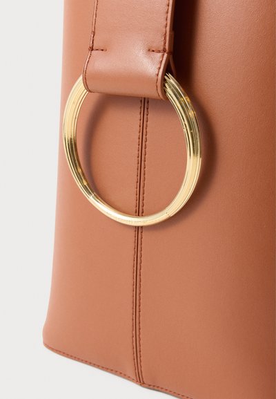 Hnedá taška z umelé kože s hladkou textúrou, ozdobená veľkým zlatým kruhovým krúžkom ako akcentom a viditeľnými detailmi švov.