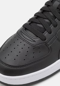 Zapatilla de cuero negra con puntera perforada, parte superior texturizada y cordones negros. Presenta una suela de goma blanca.
