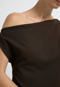 Donna che indossa un top scuro di colore marrone, con le spalle scoperte, che mostra la spalla sinistra e parte del collo e della bocca su uno sfondo chiaro e uniforme.