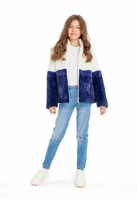 Ragazza adolescente con capelli castani mossi, indossa una giacca pelosa a blocchi di colore bianco e blu scuro, jeans azzurri chiari e sneakers bianche, in posizione rilassata.