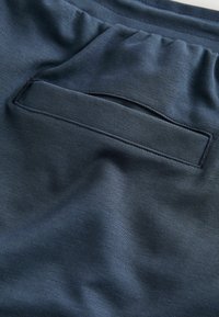 Primo piano di un tessuto blu scuro con una tasca a fessura orizzontale e cuciture visibili, che mostra la trama e lievi pieghe del tessuto.