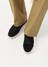 Sorte semskede sneakers med snørebånd, som har en hvid gummisål. Båret med khaki brede bukser, som viser en neutral baggrund.